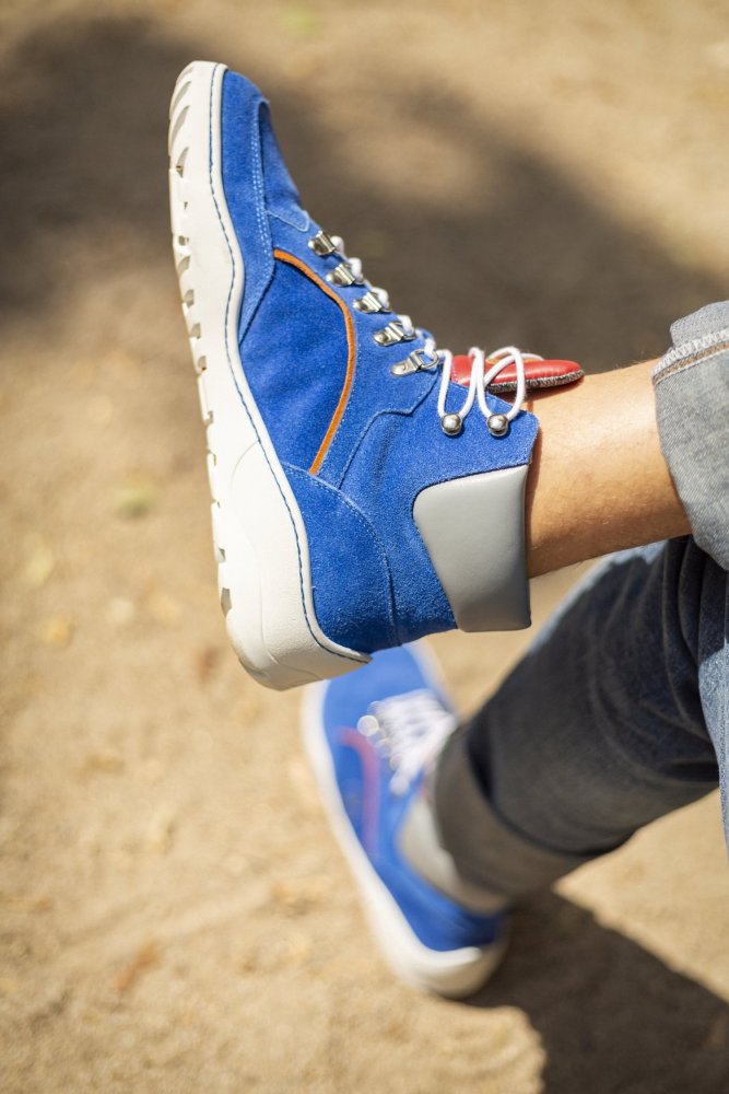 Eine Person in den stylischen TERRAQ Azure Waterproof High-Top-Sneakern von ZAQQ sitzt im Freien auf einem Sandboden. Die Schuhe aus Veloursleder haben ein blaues Obermaterial mit weißer Sohle und roten Akzenten. Silberne Ösen und Schnürsenkel vervollständigen den Look, während Jeans den lässigen Look unterstreichen.