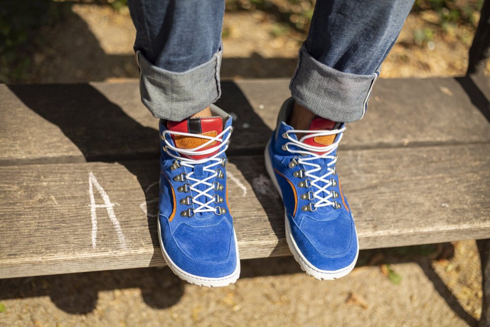 Jemand in hochgekrempelten Jeans und den blauen TERRAQ Azure Waterproof Schuhen von ZAQQ mit weißen Schnürsenkeln sitzt auf einer Holzbank. Die Schuhe haben ein leuchtend orangefarbenes Detail und das Veloursleder-Finish unterstreicht ihren stylischen Look. Kreideschrift ziert die Oberfläche der Bank und verleiht ihr zusätzlichen rustikalen Charme.