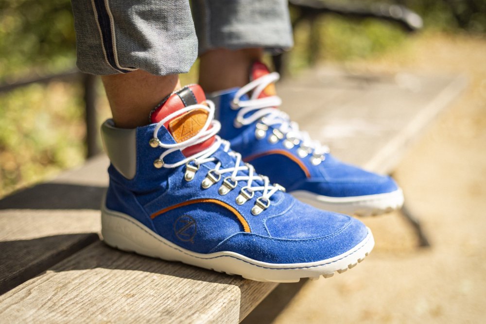 Eine Nahaufnahme einer Person, die ZAQQs wasserdichte TERRAQ Azure High-Top-Sneaker in leuchtenden Blautönen, mit weißen Schnürsenkeln und roten Akzenten trägt. Diese Schuhe aus Veloursleder sind mit Jeans mit Umschlag kombiniert, während die Person auf einer Holzbank in einer sonnigen Umgebung im Freien steht.