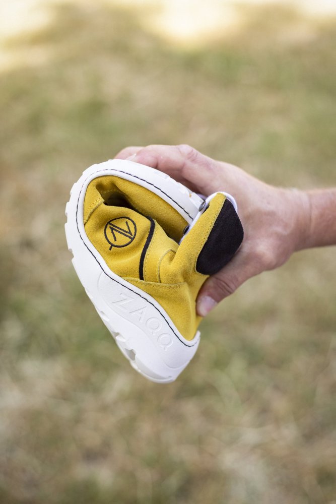 Eine Hand hält einen ZAQQ TERRAQ Yellow Waterproof Barfußschuh und biegt ihn, um seine Flexibilität zu demonstrieren. Der Schuh mit dem ZAQQ-Logo auf der Sohle scheint aus einem weichen, biegsamen Material gefertigt zu sein. Vor einer grasbewachsenen Kulisse ist er perfekt für Abenteuer im Freien.