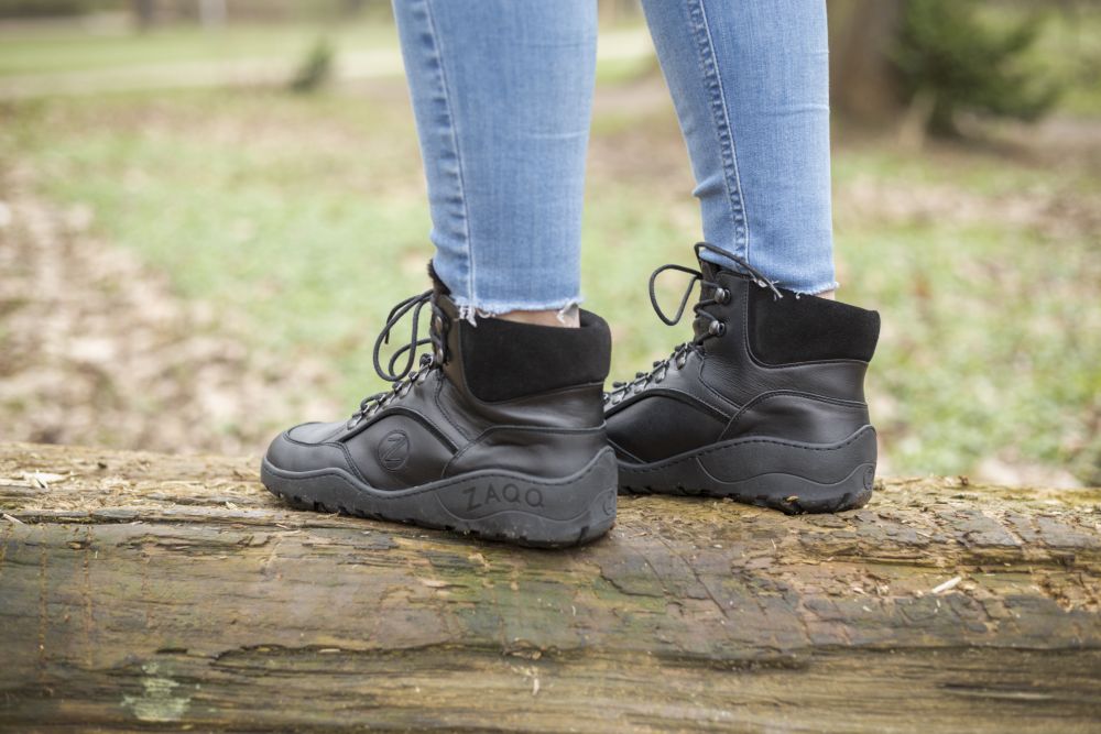 Eine Person in hellblauen Jeans und den wasserdichten TREQ Black Winter-Stiefeln von ZAQQ steht auf einem umgestürzten Baumstamm im Freien. Der Boden im Hintergrund ist mit Grün bedeckt, was die perfekte Mischung aus Abenteuer und wasserdichtem Komfort unterstreicht.