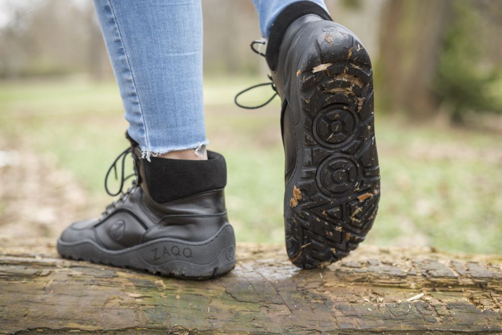 Nahaufnahme einer Person, die die wasserdichten TREQ Black Winter-Wanderstiefel von ZAQQ und Jeans trägt und im Freien auf einem Baumstamm läuft. Diese Stiefel sind ideal für Winterabenteuer und weisen ein einzigartiges Profilmuster auf, auf dessen Sohle deutlich „ZAQQ“ zu sehen ist. Im Hintergrund sind Gras und verschwommene Bäume zu sehen.