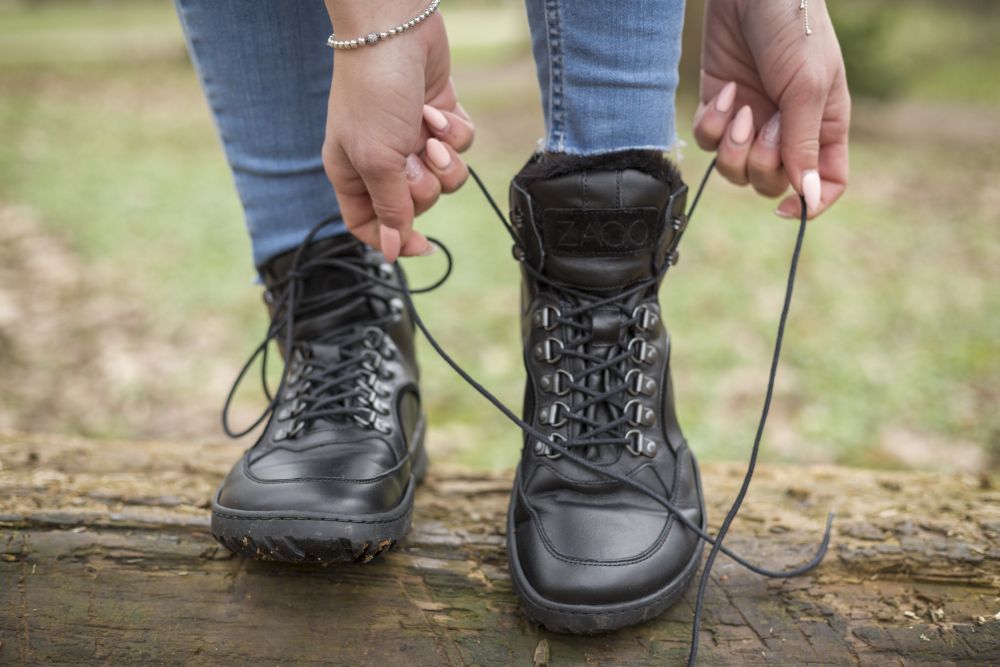 Eine Person in Blue Jeans schnürt die Schnürsenkel der wasserdichten schwarzen TREQ Winterstiefel von ZAQQ, während sie auf einem Holzstamm sitzt. Im Hintergrund ist eine grasbewachsene Außenfläche zu sehen, die sich ideal zum Testen der wasserdichten Eigenschaften eignet.