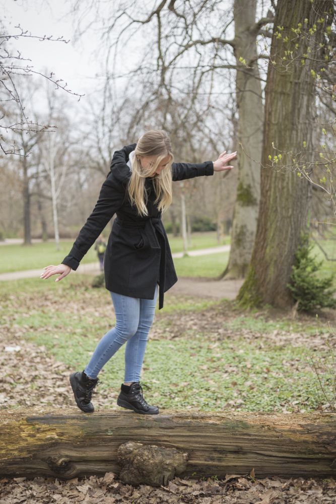 Eine Frau in schwarzem Mantel und Jeans, die schwarze, wasserdichte TREQ Winterstiefel von ZAQQ trägt, balanciert auf einem umgestürzten Baumstamm im Park. Umgeben von Bäumen und dem mit Blättern bedeckten Boden streckt sie beim Gehen ihre Arme aus, um das Gleichgewicht zu halten.