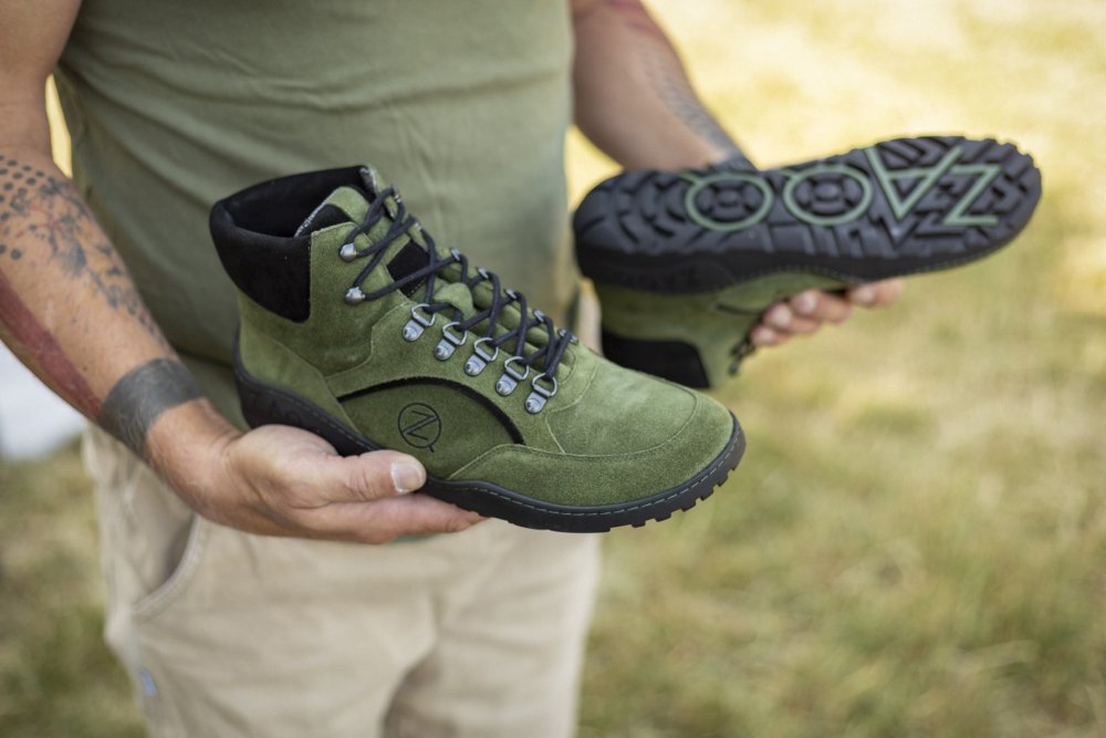 Eine Person hält ein Paar ZAQQ TERRAQ Green Waterproof Wanderschuhe mit schwarzen Sohlen. Die wasserdichten Stiefel haben Schnürsenkel und ein Logo an der Seite. Die Person trägt ein grünes Hemd und beige Hosen und zeigt Tattoos auf ihren Armen, während sie im Gras steht.