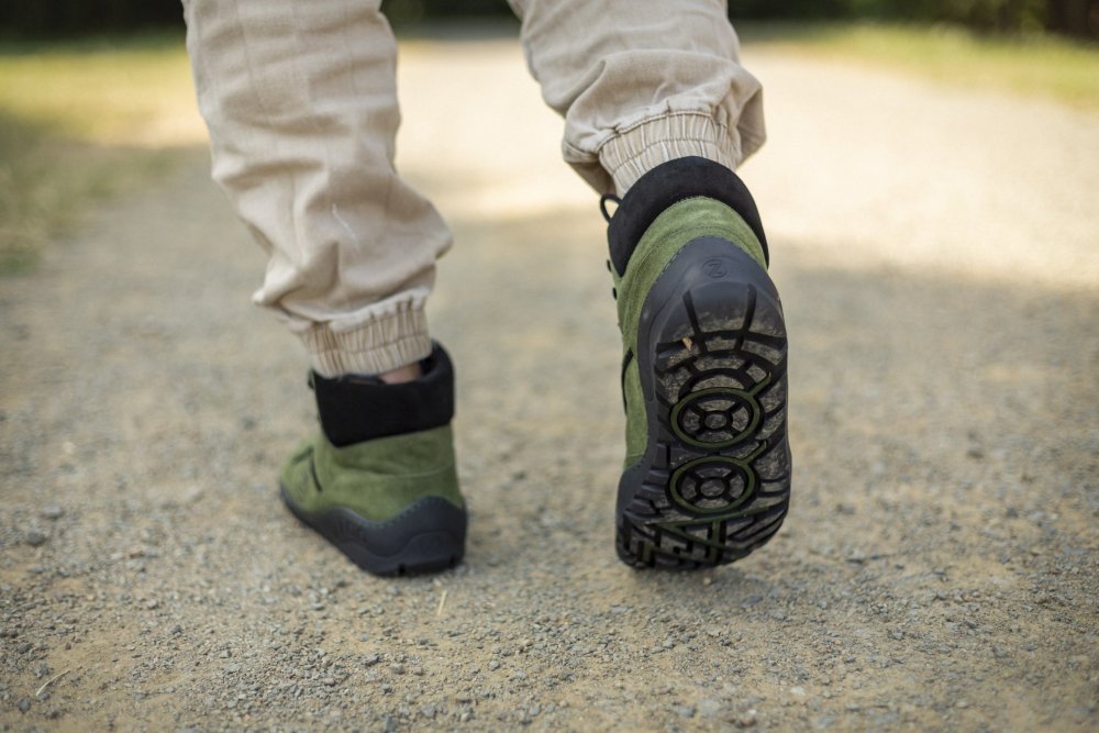 Eine Person, die die TERRAQ Green Waterproof-Stiefel von ZAQQ und beigefarbene Hosen trägt, läuft auf einem Kiesweg (von hinten gesehen). Die Sohlen der Stiefel weisen ein ausgeprägtes Profil auf, während sanftes, natürliches Licht die ruhige Außenumgebung betont.