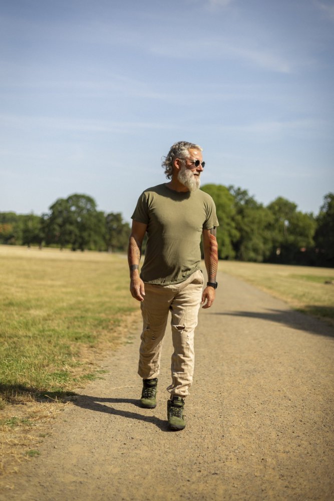 Ein Mann mit Bart und Sonnenbrille schlendert einen Kiesweg in einem Park entlang. Er trägt ein olivgrünes T-Shirt, beige Hosen und dunkle Turnschuhe. Umgeben von grünen Bäumen und Gras unter dem klaren blauen Himmel halten ihn seine grünen wasserdichten ZAQQ TERRAQ-Schuhe mit der Schönheit der Natur verbunden.