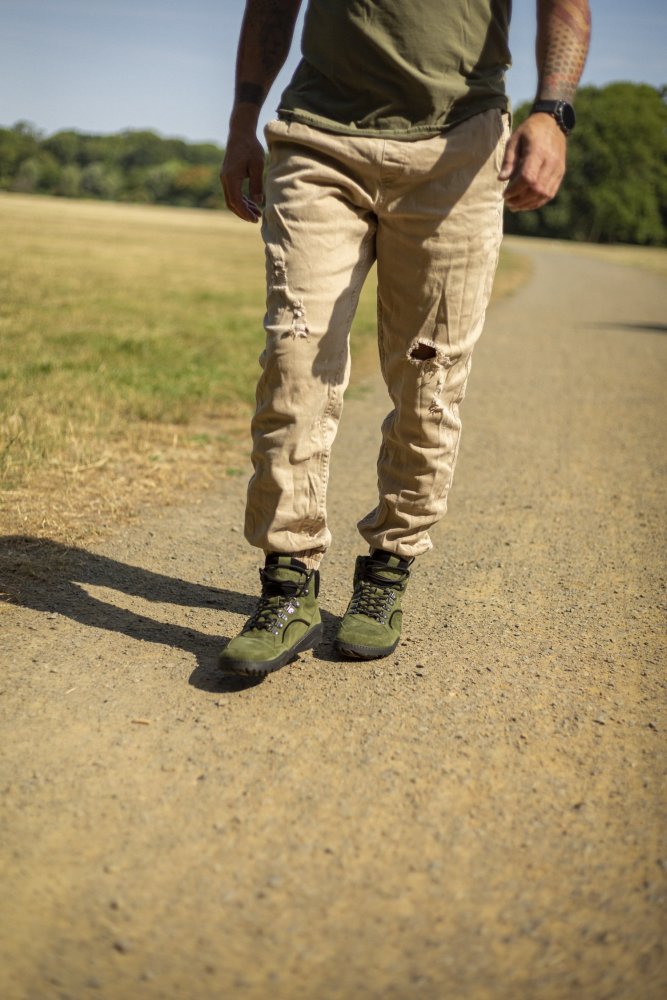 Eine Person, die in einem grünen T-Shirt, beigefarbenen Cargohosen und grünen wasserdichten ZAQQ TERRAQ-Stiefeln einen Feldweg im Park entlangspaziert. Der Weg ist von üppigem Gras und Bäumen unter einem sonnigen Himmel umgeben.