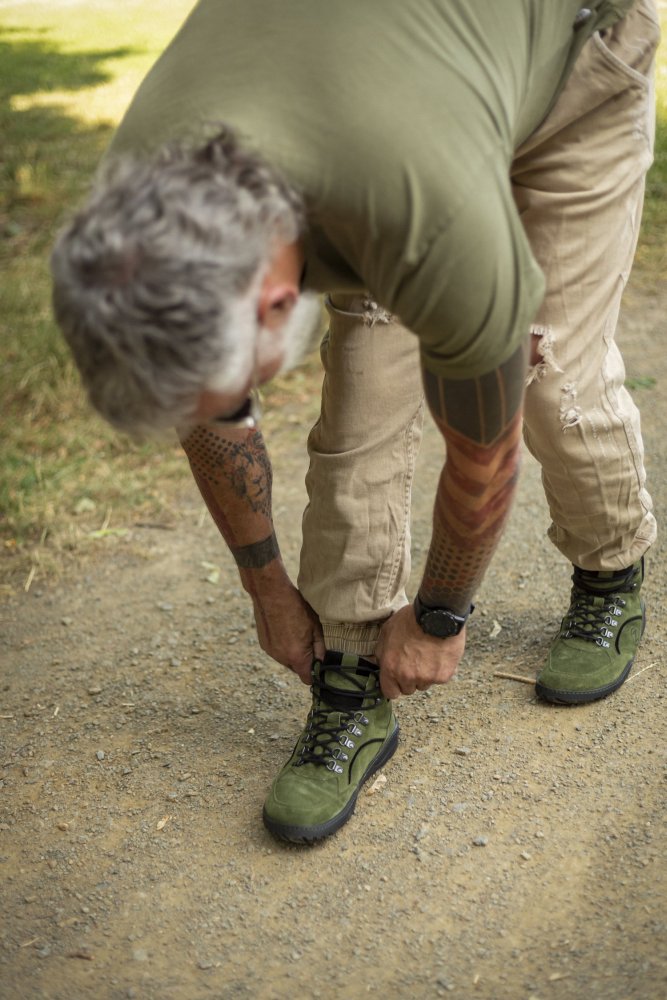 Eine Person mit grauen Haaren und Tattoos schnürt ihre grünen ZAQQ TERRAQ Green Waterproof Wanderschuhe auf einem Feldweg. Sie trägt ein grünes Hemd und eine beige Hose und genießt das Gefühl barfuß zu laufen, umgeben von der Naturkulisse mit Gras, das sich bis zum Horizont erstreckt.