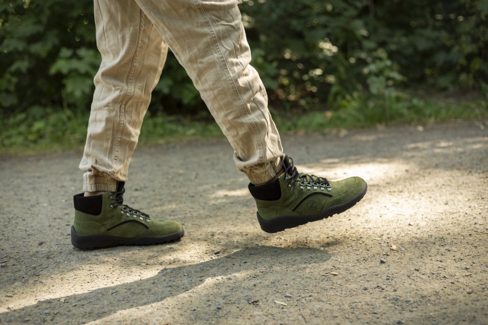 Eine Person in beigen Hosen und den wasserdichten TERRAQ Green-Wanderstiefeln von ZAQQ läuft auf einem Schotterweg durch ein Waldgebiet, umgeben von üppigem Grün.