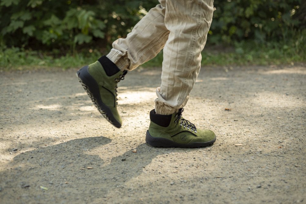 Eine Person läuft in hellbeigen Hosen und ZAQQs TERRAQ Green Waterproof Wanderschuhen aus wasserdichtem Veloursleder mit schwarzer Sohle einen Schotterweg entlang, umgeben von Grün. Der Schatten der Person ist auf dem Boden sichtbar.