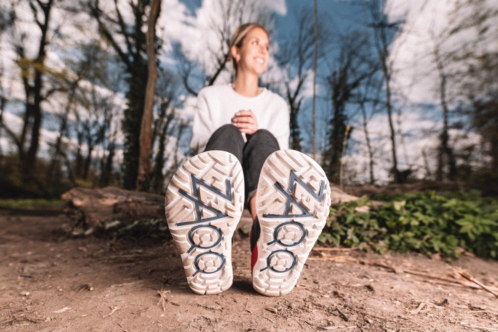 Eine Person sitzt mit ausgestreckten Beinen auf dem Waldboden und konzentriert sich auf die Sohlen ihrer Barfußschuhe. In dieser ruhigen Umgebung fällt der besondere Tragekomfort der ZAQQ VAQATION Red Waterproof auf. Bäume und Himmel lugen hervor und die Person scheint zufrieden zu lächeln.