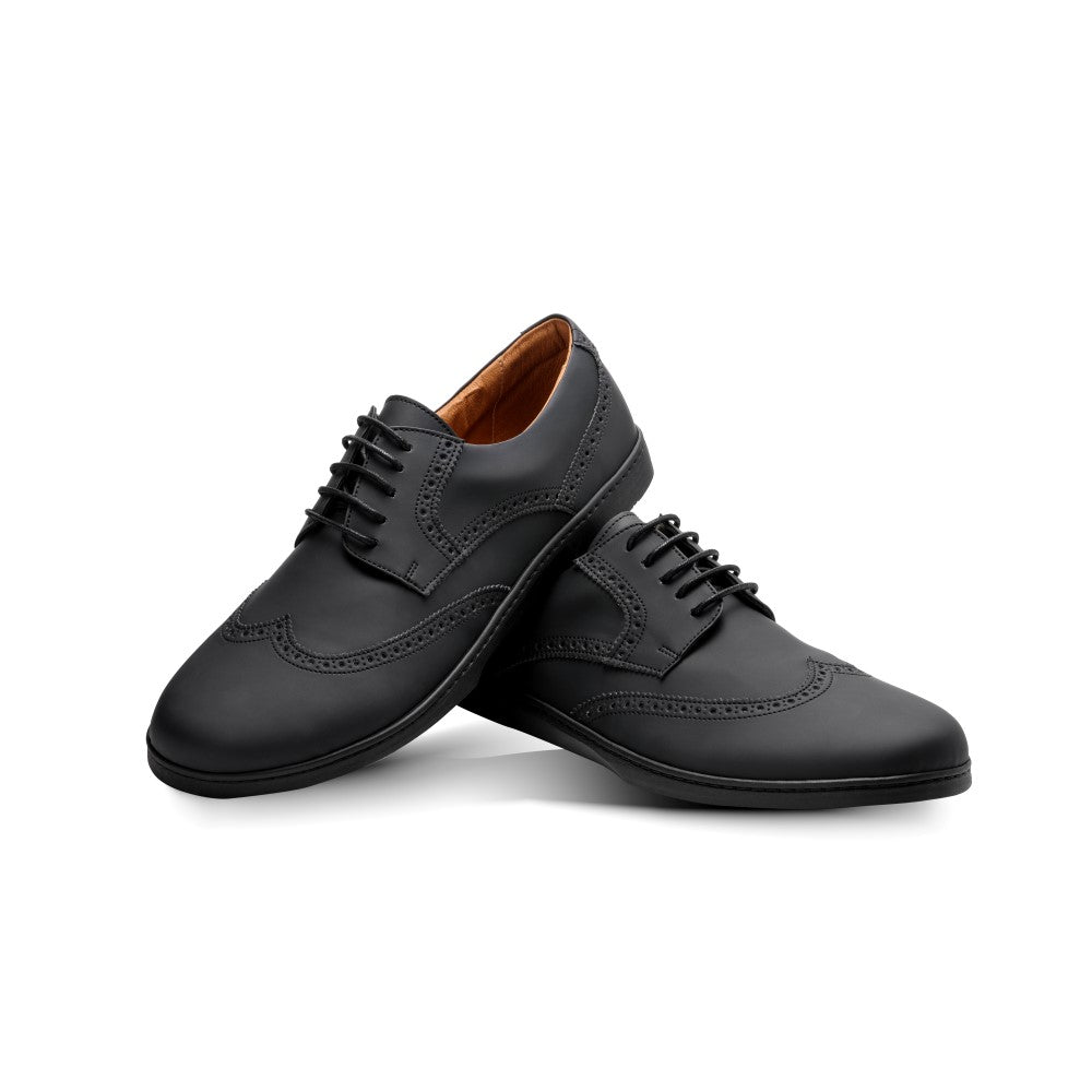 Die BRIQ Brogue Vegan Black Schuhe von ZAQQ werden auf einem weißen Hintergrund präsentiert und präsentieren ihr elegantes veganes schwarzes Design mit aufwendigen Brogue-Details und einer rutschhemmenden Sohle, sauber geschnürt, wobei ein Schuh an den anderen lehnt.