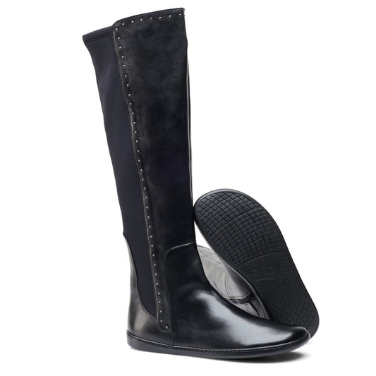 Wir präsentieren die QATCH SHINE Nappa Black-Stiefel von ZAQQ: Diese schwarzen kniehohen Stiefel haben eine glatte Lederfront und eine strukturierte Gummisohle. Das Design wird durch kleine Metallnieten an den Seiten aufgewertet und hat hinten einen dehnbaren Stoff für zusätzlichen Komfort. Ein Stiefel steht aufrecht, während ein anderer auf der Seite liegt, sodass die Sohle sichtbar ist.